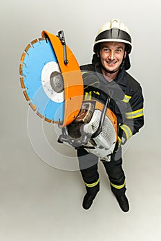 One smiling male firefighter dressed in uniform posing with saw over white studio background.