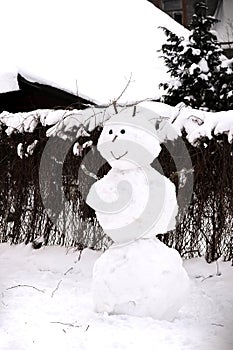 One smiling snowman with funny face standing on snow with a house and a fence covered with leafless vines of wild grapes