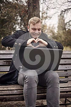 One smiling young man, sitting on bench in public park using laptop,