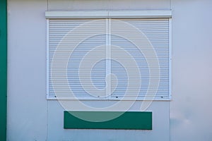 Square window closed by white roller shutters on a gray wall