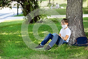 One student boy wearing mask is sitting at park with book during covid-19 pandemic