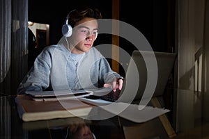 One teenager millennial using his laptop to do homework and listen music at night - online classes and lessons concept and