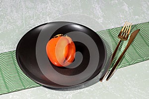 One ugly orange persimmon on black plate and golden cutlery on napkin on light backdrop.