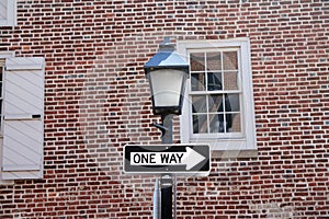 One way sign hanging on a black pole with street lamp, with a traditional brick wall on the back