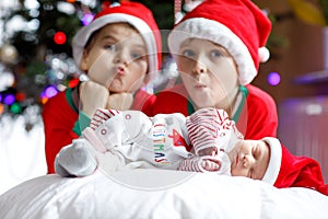 One week old newborn baby girl and two siblings kid boys in Santa Claus hats near Christmas tree with colorful garland