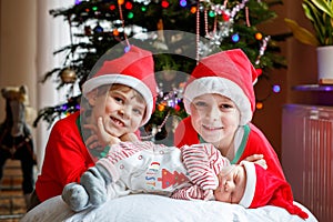 One week old newborn baby girl and two siblings kid boys in Santa Claus hats near Christmas tree with colorful garland
