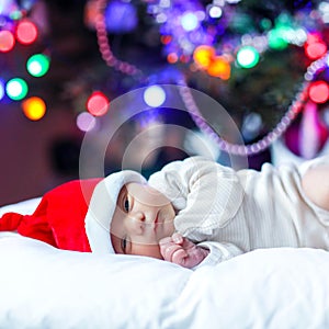 One week old newborn baby in Santa hat near Christmas tree with colorful garland lights on background. Closeup of cute