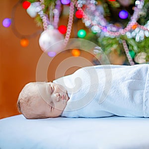 One week old newborn baby wrapped in blanket near Christmas tree with colorful garland lights on background. Closeup of