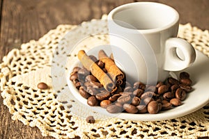 One white cup and saucer with coffee beans and a cinnamon stick stand on a wooden table in the morning