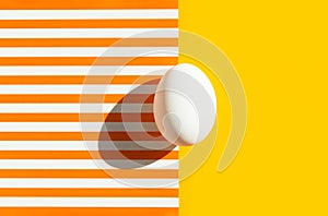 One white egg on duotone yellow orange and white striped background. Easter concept. Hard light harsh shadow. Trendy minimalist