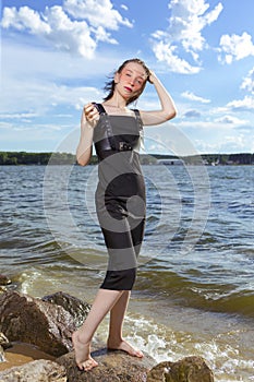 One Winsome Positive Young Happy Smiling Caucasian Brunette Girl in Black Dress With Lifted Hands At Seashore Stones During Sunny