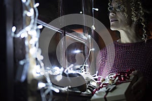 One woman at home holding christmas gift looking outside the windows with holiday lights. Happy female people waiting holiday