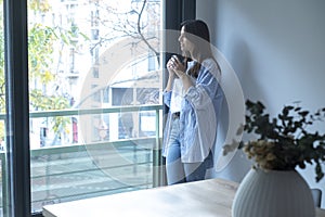 One woman at home standing and looking outside the window drinking cup of coffee and smiling. One female young adult people living