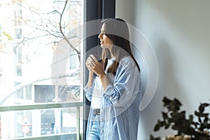 One woman at home standing and looking outside the window drinking cup of coffee and smiling. One female young adult people living