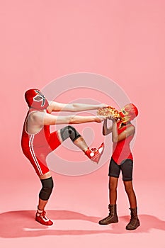 One wrestler in high kick pose holding pizza, other in mask eagerly receiving it against pink studio background.