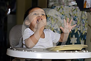 One 1 year old Asian baby boy learning to eat by himself by spoon, messy on baby dining chair at home