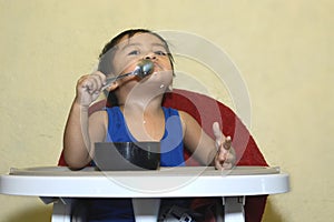 One 1 year old Asian baby boy learning to eat by himself by spoon, messy on baby dining chair