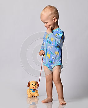 One-year-old baby boy in blue one-piece bodysuit romper with long sleeves stands with toy dog holding finger at cheek