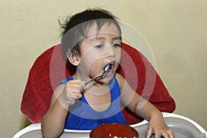 One 1 year old baby boy learning to eat alone messy on baby dining chair at home