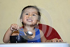 One 1 year old baby boy learning to eat alone smiling happy but messy on baby dining chair at home