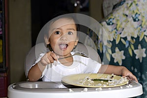 One 1 year old baby boy learning to eat alone smiling happy but messy on baby dining chair at home