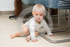 One year old baby boy sitting on floor at home and looking at camera. Little blond kid is crawling on all four under chair