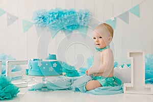 A one-year-old boy in blue shorts and a bow tie celebrates his birthday next to a cake on a stand and a decoration for his party