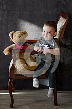 The one-year-old boy is sitting on a armchair with a bear-toy and smiles happily.