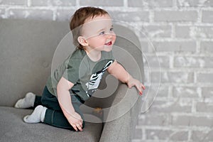 The one-year-old boy is sitting on a gray sofa and smiles happily