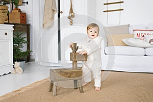 A one-year-old child in a soft jumpsuit stands next to a wooden deer toy in the living room, decorated for Christmas or New Year