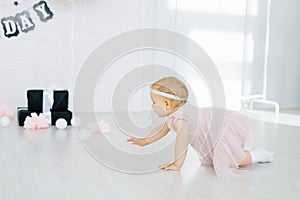 A one-year-old girl in a pink dress crawls on the floor in the children`s room