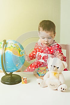 A one-year-old girl in a red dress is sitting at a table with a book, a globe and a teddy bear.