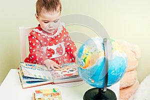 A one-year-old girl in a red dress is sitting at a table with a book, a globe and a teddy bear.