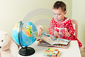 A one-year-old girl in a red dress is sitting at a table with a book, a globe and a teddy bear.