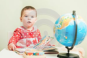 A one-year-old girl in a red dress is sitting at a table with a book, a globe and a teddy bear.