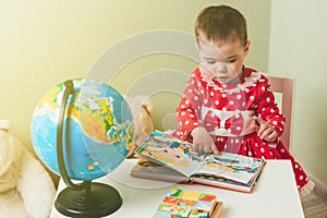 A one-year-old girl in a red dress is sitting at a table with a book, a globe and a teddy bear.