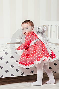 A one-year-old girl in a red dress takes her first steps holding onto the bed in the room