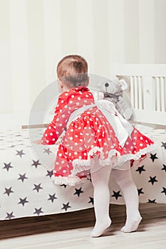 A one-year-old girl in a red dress takes her first steps holding onto the bed in the room