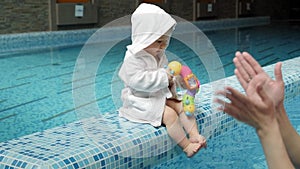 A one-year-old girl in a white coat with a toy sits on the edge of the pool.