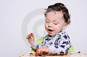 One year old kid eating a slice of birthday smash cake by himself.