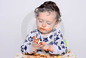 One year old kid eating a slice of birthday smash cake by himself.