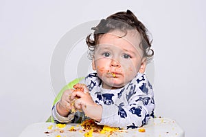 One year old kid eating a slice of birthday smash cake by himself.
