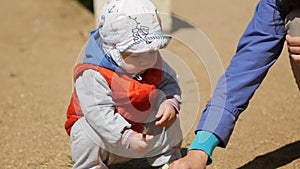 One year old kid playing with sand on the playground. Near the mother helps him with the game. Boy in the red waistcoat