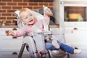 One year toddler girl sits on baby high chair with feeding bottle in her hand.