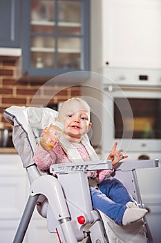 One year toddler girl sits on baby high chair with feeding bottle in her hand.