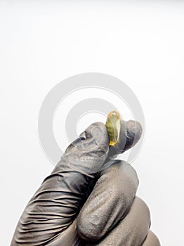 One yellow capsule of fish oil in hand in a black medical glove on a white background. Hand with pills on a white background