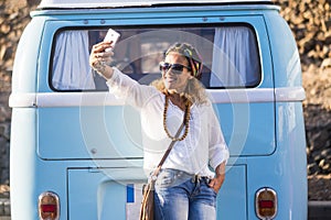 One young adult woman taking selfie picture with phone against a blue classic van. Travel and hippy lifestyle people concept. One