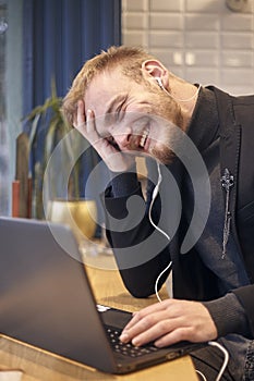 One young candidly smiling man, sitting indoors in coffee shop and using his laptop, earbuds in ear