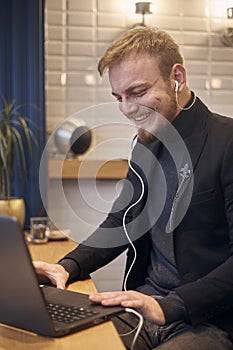 One young candidly smiling man, sitting indoors in coffee shop and using his laptop, earbuds in ear