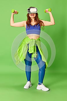 One young charming girl, woman wearing vr glasses doing workout with dumbbells and smiling at camera over green